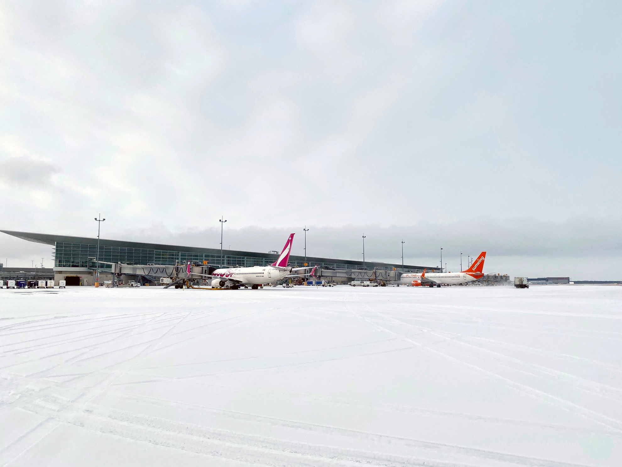 Un avion Swoop et un avion Sunwing sont stationnés aux passerelles d'embarquement de l'aéroport international Richardson de Winnipeg, entourés d'une aire de trafic enneigée.