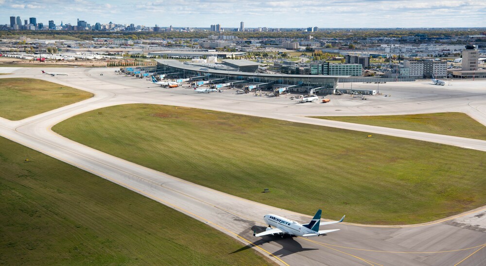 A WestJet plane taxis towards the Winnipeg Richardson International Airport terminal with the city skyline in the background.