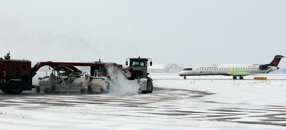 Un chasse-neige déblaie la neige sur le terrain d'aviation alors qu'un avion d'Air Canada roule en arrière-plan.